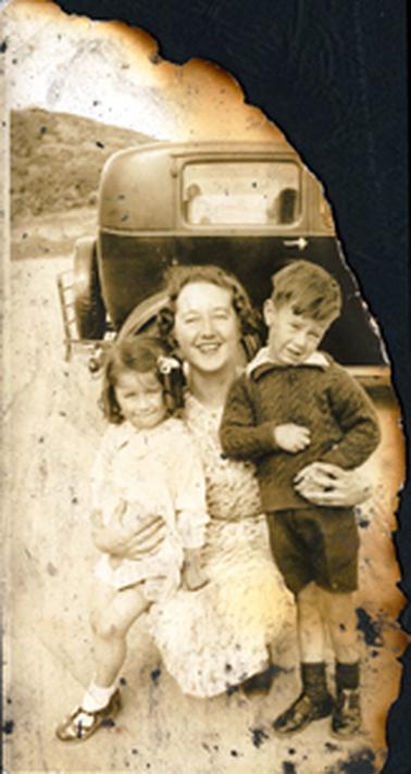 This badly damaged image shows a woman squatting on the ground with her right arm around a little girl and her left arm around a small boy.  The woman has curly hair and is wearing a print dress.  The little girl has dark coloured ringlets, long sleeves and white socks and black shoes.  The small boy is wearing a white shirt under his cable knit pullover, dark coloured socks and black shoes.  Part of a motor car can be seen behind the group and a hill is in the background.  