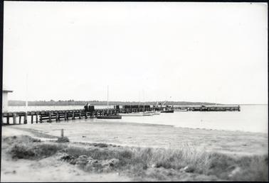 The image depicts the Hastings Pier with several wooden boats moored to the pier. The sandy foreshore is covered with seaweed. The fishing shed is located on the left and can only just be seen. 