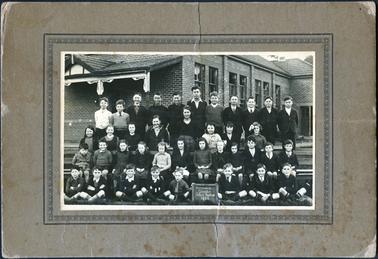 A group of 39 school children ranging in ages are standing and seated outside a brick school building. The building is the Hastings Catholic School known as the Immaculate Conception School.