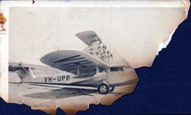 This badly torn and burnt photograph shows the wings and back section of an aeroplane.  Printed underneath the wing in very large letters is: “VH-”.  Printed on the body of the plane is: “VH-UPB”.