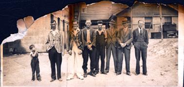 This badly burnt and torn image shows the bottom half of a photograph.  There are seven men standing in a line posing for a photo and a small boy on the left-hand side is looking at the group. The two men at the left are wearing cricket blazers and the three men on the right are wearing three-piece suits.  A group of buildings are behind the group.  