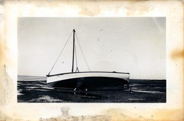 This black and white image shows a foreshore at low tide with a boat sitting on the sand.  The bottom part of the hull is black with a white band above it.  It has one mast with three support lines.  The rope from the anchor can be seen on the left-hand side.  The very small printing on the side could read: AF61048 2636.  