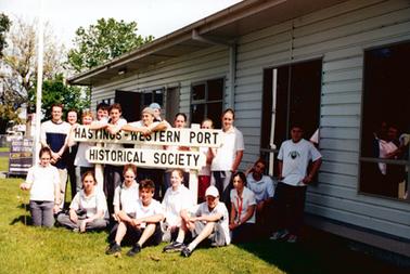 This image shows a group of nineteen young people standing and sitting on the grass outside the Hastings-Western Port Historical Society building.  Two of the boys have their arms folded on the top of the Museum sign.  A board which reads “Historical Museum Now Open” is to the left of the group.  
