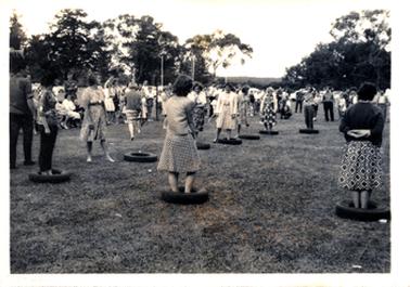 This image shows a large group of people enjoying a social occasion.  At the centre of the image, approximately eight woman are standing inside car tyres.  The two women in the foreground have their backs to the camera.  All except two of the women are wearing dresses and some are also wearing cardigans.  A large group of men, women and children are standing and sitting around watching.  Tall trees are in the background.   