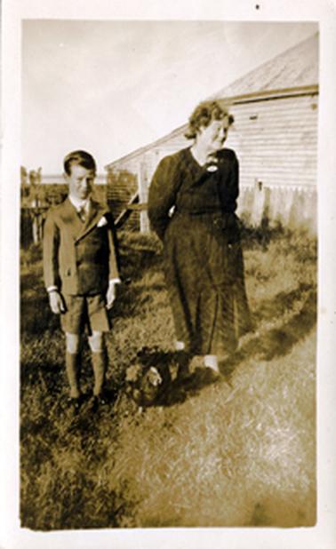 This image shows an elderly woman and a young boy standing side by side, posing for the camera.  A hen is sitting on the grass between the two.  The woman is wearing a long sleeved dark coloured dress with a brooch at the neckline.  The boy is wearing a white shirt, tie and double breasted suit and long socks.  Part of a weatherboard house can be seen on the right-hand side and water can be seen in the far distance.  