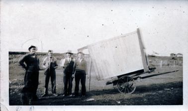 This image shows four men standing facing the camera beside a two-wheeled covered ‘box’ wagon in an open field.  Three of the men are wearing hats.  A fence can be seen in the distance in front of a paddock.   