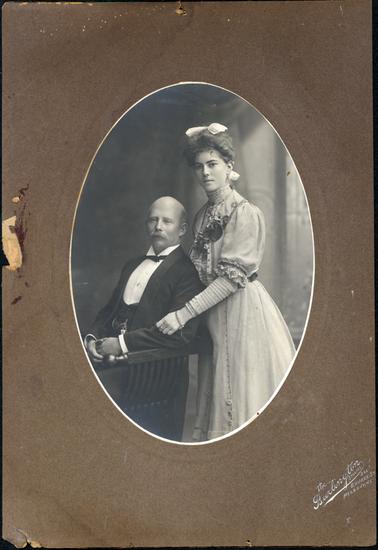 This is an oval portrait of a beautifully dressed man and woman, posing for a photograph.  They are both turned slightly to the side.  The man is sitting in a wooden arm chair and is dressed in evening clothes.  He has a watch and chain attached to his waistcoat.  He has a bald head and a grey moustache.  The young woman is standing behind the chair and her left hand is resting on the man’s arm.  Her dress has elaborate pin tucks and frills.  She has pearls above her very high neckline and a spray of silk flowers on the left-hand side of the bodice.  He hair is piled high with a large white bow at the top and at the back.  Written on the bottom of the mounting in silver ink is: “The Burlington Studios 294 Bourke St. Melbourne”.