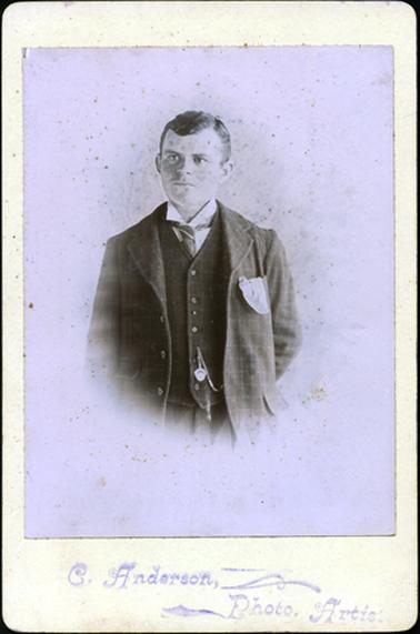 This portrait shows a young man, taken from the waist up.  He is dressed in a three piece check suit, white shirt and striped tie.  His hair is neatly parted on the left side.  He has a watch and chain attached to his waistcoat and a handkerchief hanging out of his left coat pocket.  He is looking away from the camera.  Printed on the mounting at the bottom is: “C. Anderson, Photo Artist”.