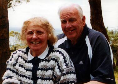 This image shows the top half of a man and woman with broad smiles for the camera.  She has fair hair and glasses and the man has grey hair.  They are both wearing jumpers.  Two large tree trunks are behind the couple and small trees are in the background.  