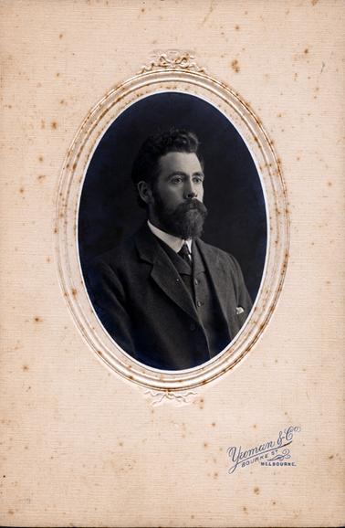 This is a portrait of a young man, taken from the chest up.  He is dressed in a dark coloured three piece suit, white shirt and tie.  He has a mop of dark wavy hair, side burns and a thick moustache and beard.  He is turned slightly to the side and is staring away from the camera.  The photo is oval shaped and has an ornate mounting.  The photographers details are stamped in blue at the bottom: ‘Yeoman & Co Bourke St. Melbourne’.