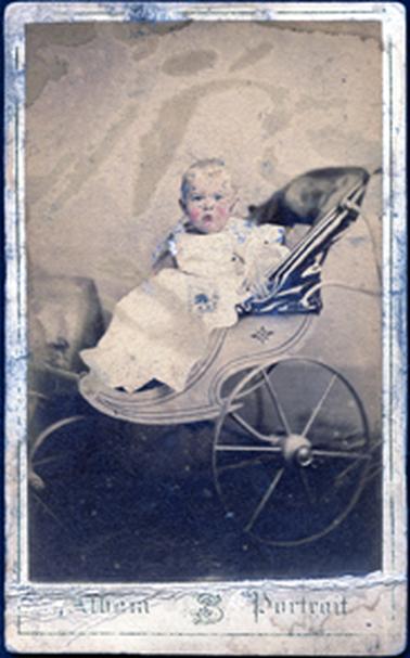 This is a studio portrait of a baby sitting up in a stroller, looking at the camera.  The baby is wearing a long white dress.  The top of the troller is partly folded back and there is a pattern on the side of the stroller.  There are two very large back wheels and one small wheel at the front.  The following is printed across the bottom mounting: “Atham  S  Portrait”.  