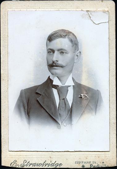 This portrait shows a young man, taken from the chest up.  He is dressed in a three piece suit, white shirt and tie.  His hair is neatly parted on the left side.  He has a flower in his coat lapel.  He has a large moustache and he looking away from the camera.  The photographer’s details are printed on the bottom of the mounting - ‘E. Strawbridge 129 Park St., Parkville’.