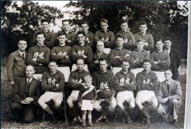 This image shows a football team standing and sitting in three rows, posing for a photo.  There are six men standing in the back row, nine men standing in the middle row and seven men sitting and kneeling in the front row.  A small boy is standing in front of the team.  The players are wearing long sleeved dark coloured shirts and white shorts.  They have the initials HFC on the front of their shirts.  Four of the non-playing men are wearing suits.  Tall trees are in the background.  