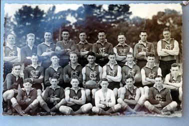 This image shows a football team standing and sitting in three rows, posing for a photo.  There are nine men standing in the back row, nine men in the middle row and six men sitting cross-legged on the ground.  Their football shirts have the initials SFC on the front.  The player at the centre of the middle row is holding a football.  Tall trees are in the background.  