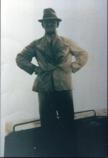 This image shows a man standing at the back of a boat with his hands on his hips.  He is wearing dark coloured pants, a light coloured jacket and hat.  He is wearing glasses.  Water can be seen behind him. 