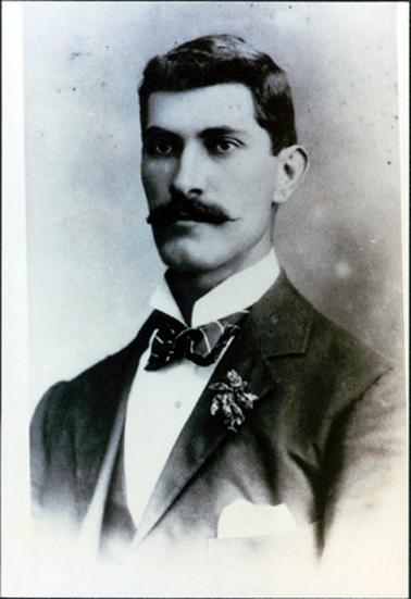 This image is a portrait of a man, taken from the chest up.  His body is turned slightly to the side.  He is wearing a three-piece suit.  His white shirt has a stand up collar.  He has a striped bow tie, a spray of flowers in his buttonhole and a white handkerchief in his coat pocket.  His black hair is parted on the left-hand side and he has a large black moustache. 