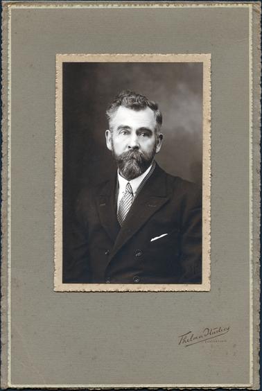 This is a studio portrait of a man, taken from the waist up.  He is wearing a dark coloured, double breasted suit, white shirt and striped tie.  A white handkerchief is in his left pocket.  He has thick, wavy partly grey hair, beard and moustache.  The photographers details are stamped on the mounting.  