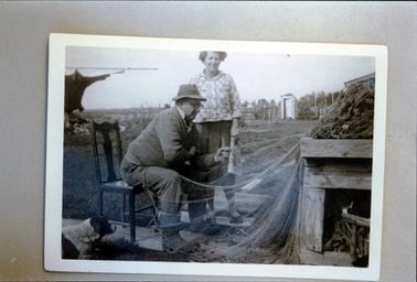 This image shows a side view of a man sitting on a wooden dining-room chair, mending fishing nets.  A woman holding a newspaper in her left hand, is standing beside him, smiling at the camera.  Nets are piled high on a wooden bench in front of the man.  A dog is sitting to the right of the man.  Clothes can be seen on a line at the far left-hand side of the photo and a wooden toilet is in front of the back wooden fence.  Tall trees can be seen in the background.  