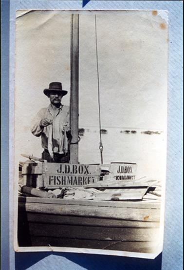 This image shows a small section of a wooden boat with a man standing beside the mast holding a fish in each hand.  He is wearing dark coloured trousers, a light coloured open neck shirt and a hat.  He has a large moustache.  Two wooden boxes have ‘J.D. Box. Fishmarket’ printed on the side. 