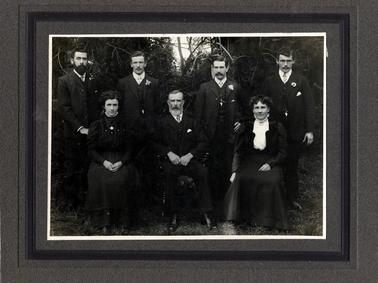 This image shows a family portrait.  There are four young men standing and one young woman and a middle-aged man and woman sitting on chairs.  They are all beautifully dressed in black.  All the men have moustaches.  The man seated and the young man at the back, left-hand side, both have beards.  All the young men have a flower in their coat lapel.  The women both have very high necklines.  Grass is in the foreground and trees are behind the group.   