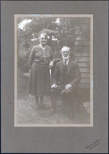 This portrait shows an elderly man sitting on a dining-room chair and a woman standing beside him, her right arm resting on the chair, posing for a professional photograph.  The woman is wearing a knee-length, long sleeved dress with a belt at the waist.  She has a string of pears tied around her neck.  The man is dressed in a three-piece suit, white shirt and tie.  Part of a weatherboard house and trees and shrubs can be seen behind them.  The photographers details are printed on the mounting.  