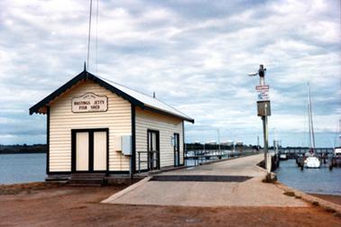 This is a photograph of the Hastings fishing shed with the jetty to the right.  The sign on the end of the building reads: “Circa 1866 Hastings Jetty Fish Shed”.  A post is on the right-hand side of the jetty with a caution sign and a street light.  Boats can be seen on either side of the jetty and large tanks are in the background.  
