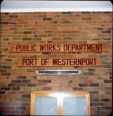 This image shows two sign boards attached to a brick wall.  They read: “PUBLIC WORKS DEPARTMENT” and “PORT OF WESTERNPORT”.  A bar heater is attached to the wall underneath the signs and part of a two-door cupboard is at the bottom of the image.  