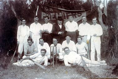 This image shows a cricket team standing, sitting and lying, posing for a photo.  There are seven men standing in the back row, five men squatting in the middle row and two men half-lying on the grass at the front.  An elderly man, dressed in a three piece suit and hat, is in the middle of the back row.  Cricket stumps are in the ground between the two front players.  They both have a cricket bat lying across their body.  The two men on either end of the back row are holding a cricket bat.  A banner with ?.C.A. Premiers is behind the group.  Trees are in the background.  