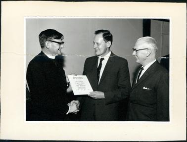 This image shows three men dressed in suits.  The two men on the left are shaking hands and holding a document which reads: “Hastings and District Bush Nursing Hospital This is to certify that Dr John F. Williams was elected a Life Governor of Hastings and District Bush Nursing Hospital on 7th day of June 1968”. A grey-haired man on the right-hand side is watching the presentation.  