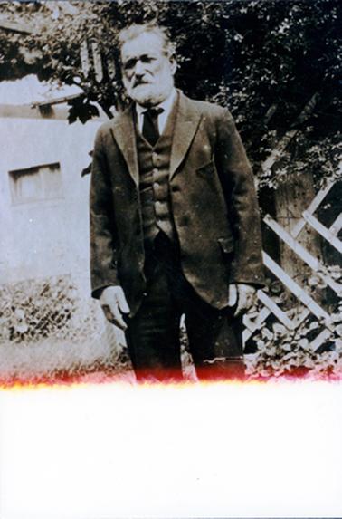 This image shows an elderly man, from the knees up, posing for a photo.  He is wearing a three piece suit, white shirt and dark coloured tie.  He has grey hair and beard and has a serious look on his face.  A fence and tree is behind him.  