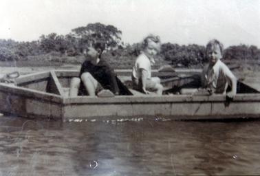 This image shows three small children in a boat close to the shoreline.  Trees and shrubs are in the background.  The two boys in the light coloured shirts are looking at the camera.  The boy on the left, in the dark coloured long-sleeved shirt is looking away.  