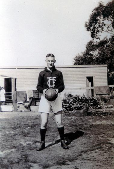 This image shows a young man, dressed in a football uniform posing for a photo.  He is dressed in a long-sleeved dark coloured shirt, white shorts and long black socks.  White coloured initials are on the front of his shirt.  He is standing with feet apart holding a football.  A pile of wood is behind him to the right and a woman is standing outside a timber building on the far left-hand side.  