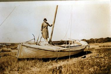 This image shows a woman standing on the top of a wooden fishing boat smiling for the camera.  Her left hand is resting on a pole.  She is dressed in a knee-length, long-sleeved dress.  The boat is on dry land with grass around it.  