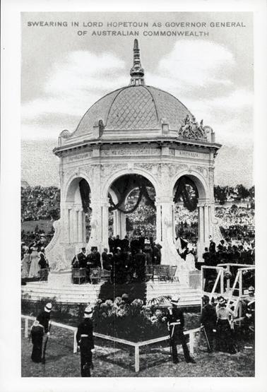 This image shows a ceremony taking place in an open white pavilion.  Decorations can be seen inside the pavilion.  A large crowd is watching.  Three policemen are in the foreground behind a white post and rail fence.  The following is printed across the top of the photograph: “Swearing in Lord Hopetoun as Governor General of Australian Commonwealth”.