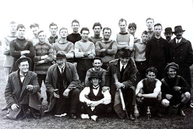 This image shows a group of twenty-four men sitting and standing, posing for a photo.  There are seventeen men standing and seven men sitting and kneeling.  Some of the men are wearing football uniforms and the rest suits, coats and hats.  The man at the front, third from the right, is holding a rolled-up banner in his left hand.  