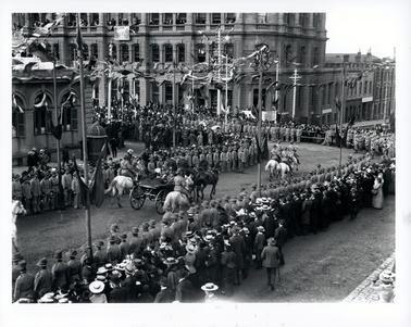 This image shows a street parade.  The streets are lined with military men and the public behind them.  People are also on the verandahs and windows of the buildings.  Soldiers on white horses are escorting a carriage drawn by two black horses.  Flags decorate the streets and buildings.  