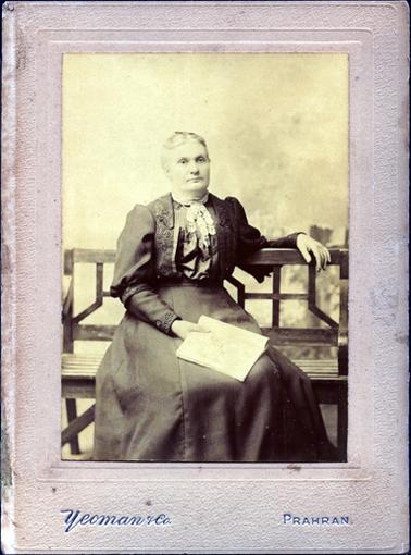 This image shows a woman sitting on a wooden bench seat.  She is holding a book in her right hand and her left arm is draped over the back of the chair.  She has a plain skirt and an embroiled jacket.  Lace is tied at the high neck line.  Her hair has a center part and is pulled back severely.  Printed in silver across the bottom of the mounting board is: “Yeoman & Co.” and ‘Prahran’.