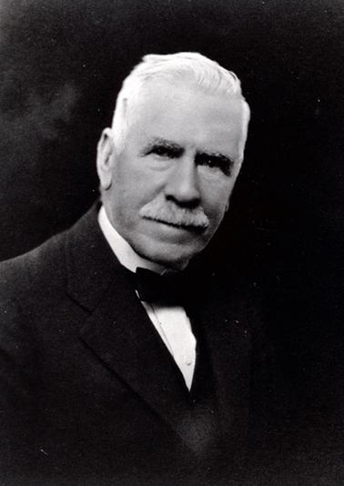This image shows a portrait of a man, taken from the waist up.  He has grey hair and a grey moustache.  He is wearing a three piece suit, white shirt and a black bow tie.  
