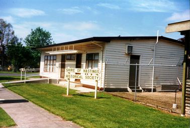 This image shows a weather board building with a flat roof.  There are two front doors and a set of windows on either side of the doors.  A concrete ramp with iron rails lead up to the left-hand side door.  Another door, with two steps, is on the side of the building.  A tall wire fence stretches from the corner of the building to another structure.  A sign on the grass beside the footpath reads: “Shire of Hastings Historical Society”.  