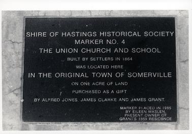 This image shows a metal plaque attached to stone.  The plaque reads: “Shire of Hastings Historical Society Marker No. 4 The Union Church and School built by settlers in 1864 was located here in the original town of Somerville on one acre of land purchased as a gift by Alfred Jones, James Clarke and James Grant.  Marker placed in 1985 by Eileen Maslen, present owner of Grant’s 1859 residence”.