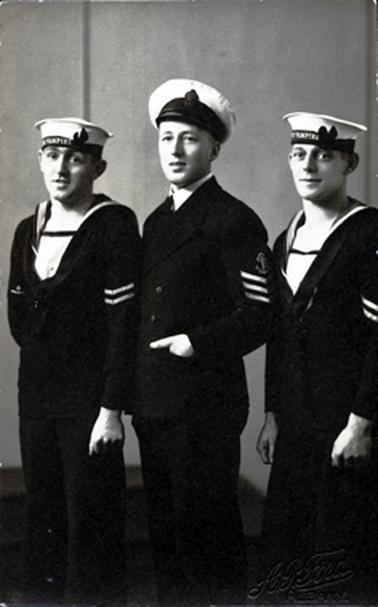 This image shows three sailors standing, slightly side-on, posing for a photo.  They are all dressed in navy uniforms and the man in the middle is dressed in an officers uniform.  The two sailors have HMAS Vampire printed on their caps.  The officer has his left hand in his coat pocket.  The photographers details are embossed at the bottom right-hand corner. 