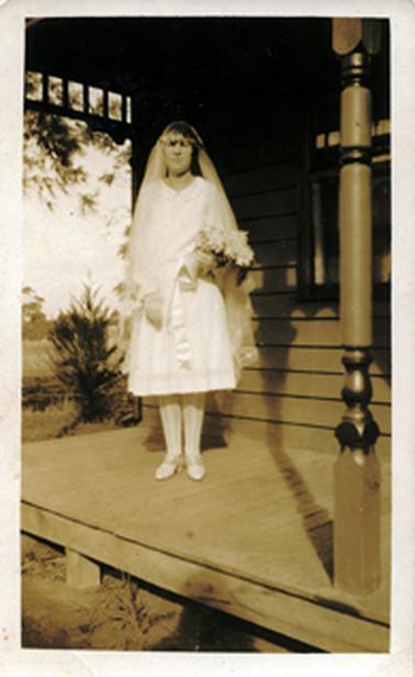 This image shows a bride standing on the verandah of a weatherboard cottage, posing for a photo.  Her knee-length white wedding dress has long sleeves and a round collar.  Her veil is attached to a floral headband and is the same length as the dress.  She is holding a white bouquet with long satin ribbons.  She is wearing white shoes and stockings.  A turned verandah post is on the right-hand side and a window is behind her.  