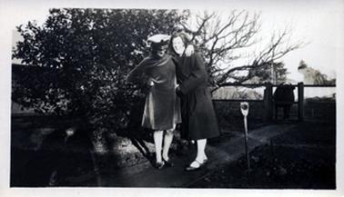 This image shows two women standing on a garden path.  They are both laughing.  Both women are wearing dark coloured coats and the woman on the left is wearing a naval cap.  She has her left arm around the neck of the other woman.  A cow is behind the couple, looking through the gate.  A garden fork is standing in the garden on the right-hand side and a tree is behind them.   