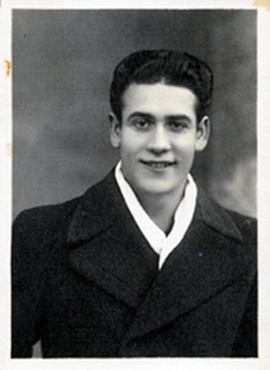 This image shows a portrait of a young man, taken from the chest up.  He is looking at the camera and has a broad smile on his face.  He has a thick head of neatly combed black hair.  He is wearing a heavy black overcoat and has a white scarf around his neck.  He is of European appearance.  There are two rust marks on the left-hand border.  