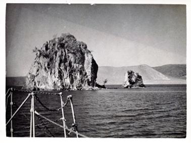 This image shows a seascape which features a very large rock and a smaller one to its right.  Water surrounds the rocks and hills can be seen in the distance.  Poles and chains are in the foreground on the far left-hand side of the image.  