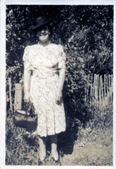 This faded image shows a woman standing in front of a fence, posing for a photo.  She is wearing a knee-length floral dress with elbow length sleeves.  She has a black hat, tilted to the side and is holding a handbag and gloves in her right hand.  She is wearing white earrings and necklace.  Untidy grass is all around her and shrubs are behind the fence.  
