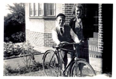 This image shows a woman and a boy standing in the front yard of a house, posing for a photo.  They are both smiling.  The boy is holding a bicycle.  He is dressed in a long-sleeved white shirt, tie, vest, shorts, long socks and a cap.  He is wearing spectacles.  The woman is wearing a plain coloured, knee length dress and a dark coloured cardigan.  The brick and concrete house is behind the couple.  A garden and path is on the left-hand side of the image.  