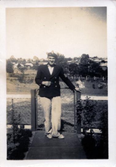 This image shows a young man standing on a garden path, posing for a photo.  He is dressed in a ‘navy outfit’, dark coloured double-breasted coat, white shirt, trousers and shoes and a cap.  A post and wire fence is behind him and his left hand is resting on the gate post.  Garden beds are on either side of the path and a town can be seen in the background.