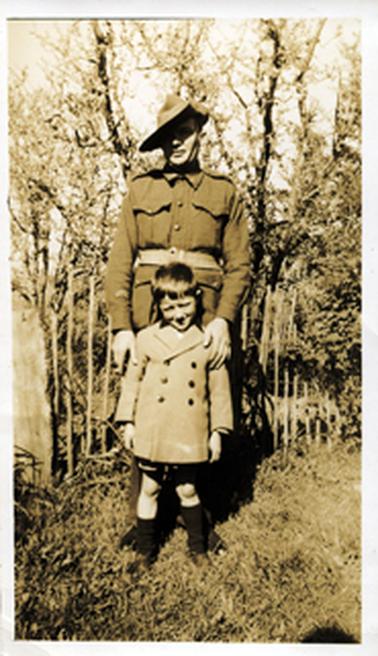 This faded image shows a man in soldier’s uniform standing in front of a fence with a small boy in front of him, posing for a photo.  His hands are resting on the top of the boy’s arms. The boy is wearing a double-breasted coat and long black socks.  They are standing in untidy grass and trees are behind the fence.  