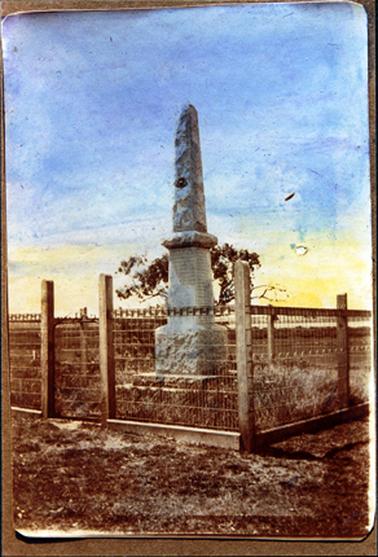 This image shows a monument standing on open ground surrounded by a post and wire fence. A tree can be seen behind the memorial.   
